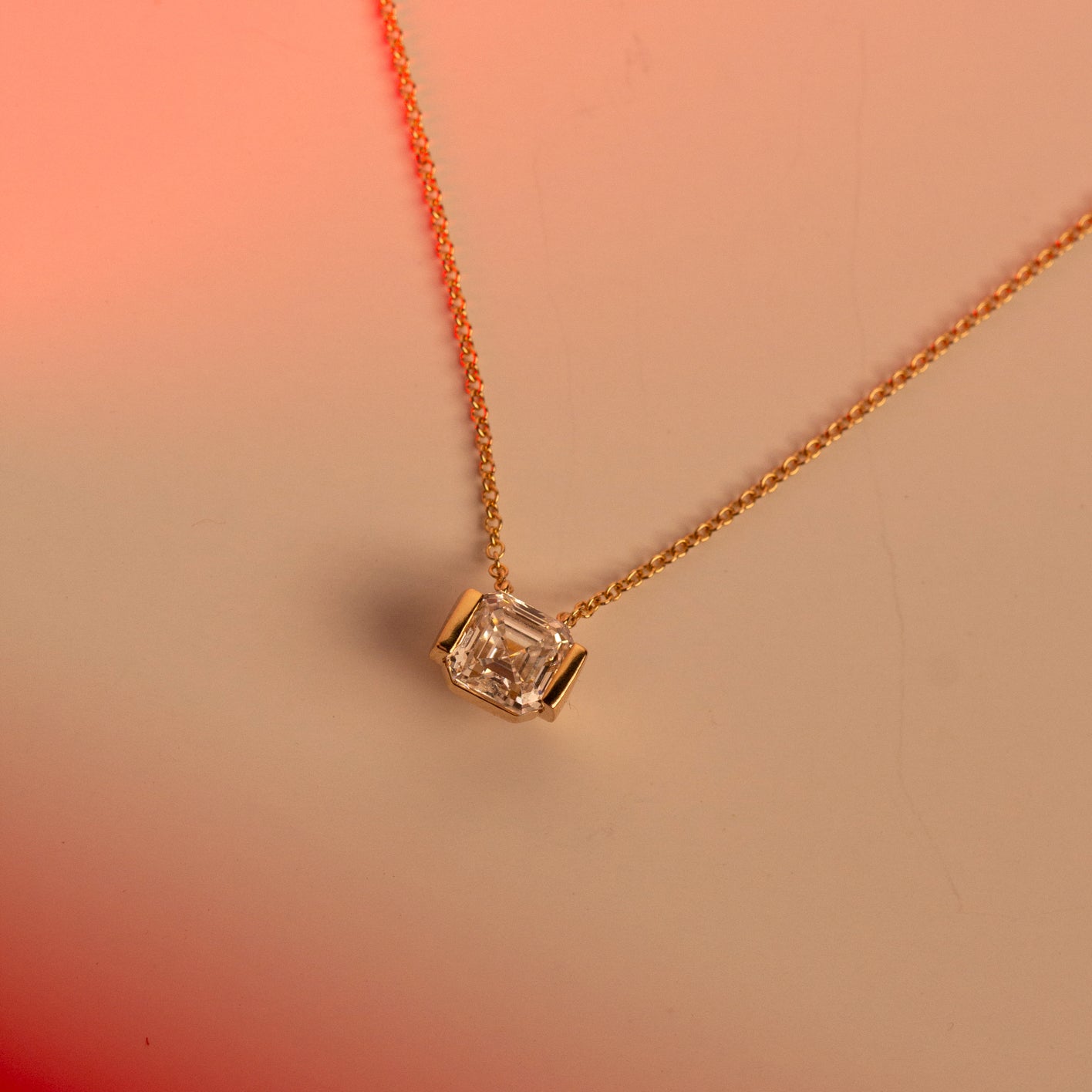 A delicate gold chain necklace with a single asscher-cut lab-grown diamond pendant in a gold half-bezel setting, displayed on a warm peach and pink gradient background.